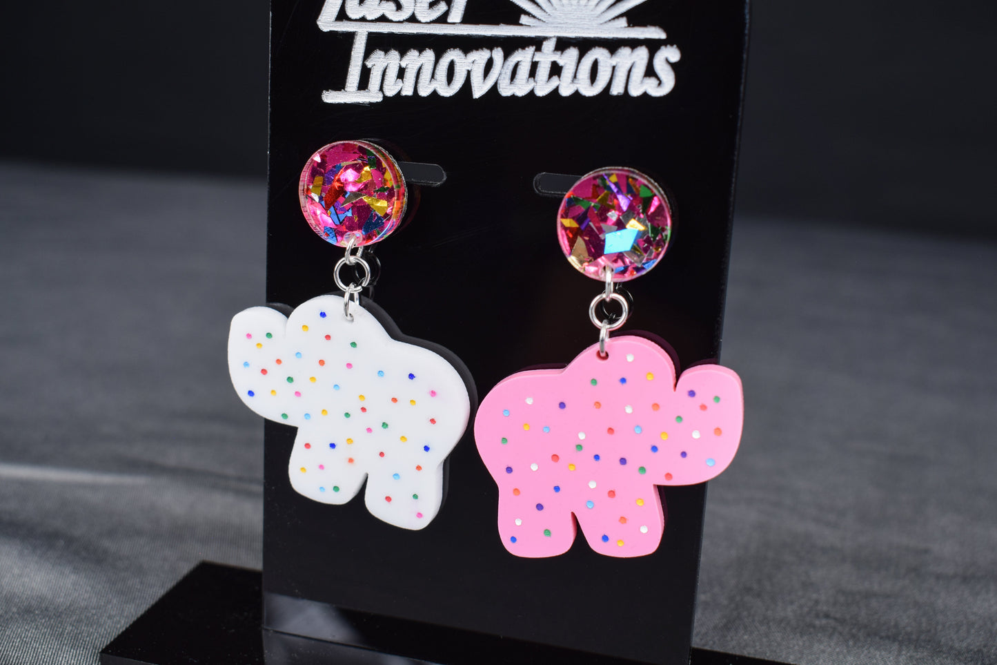 A pair of earrings featuring a white acrylic cookie-shaped elephant and a pink acrylic cookie-shaped camel, with a glittery multicolored acrylic circle attached to each and hand-painted dots