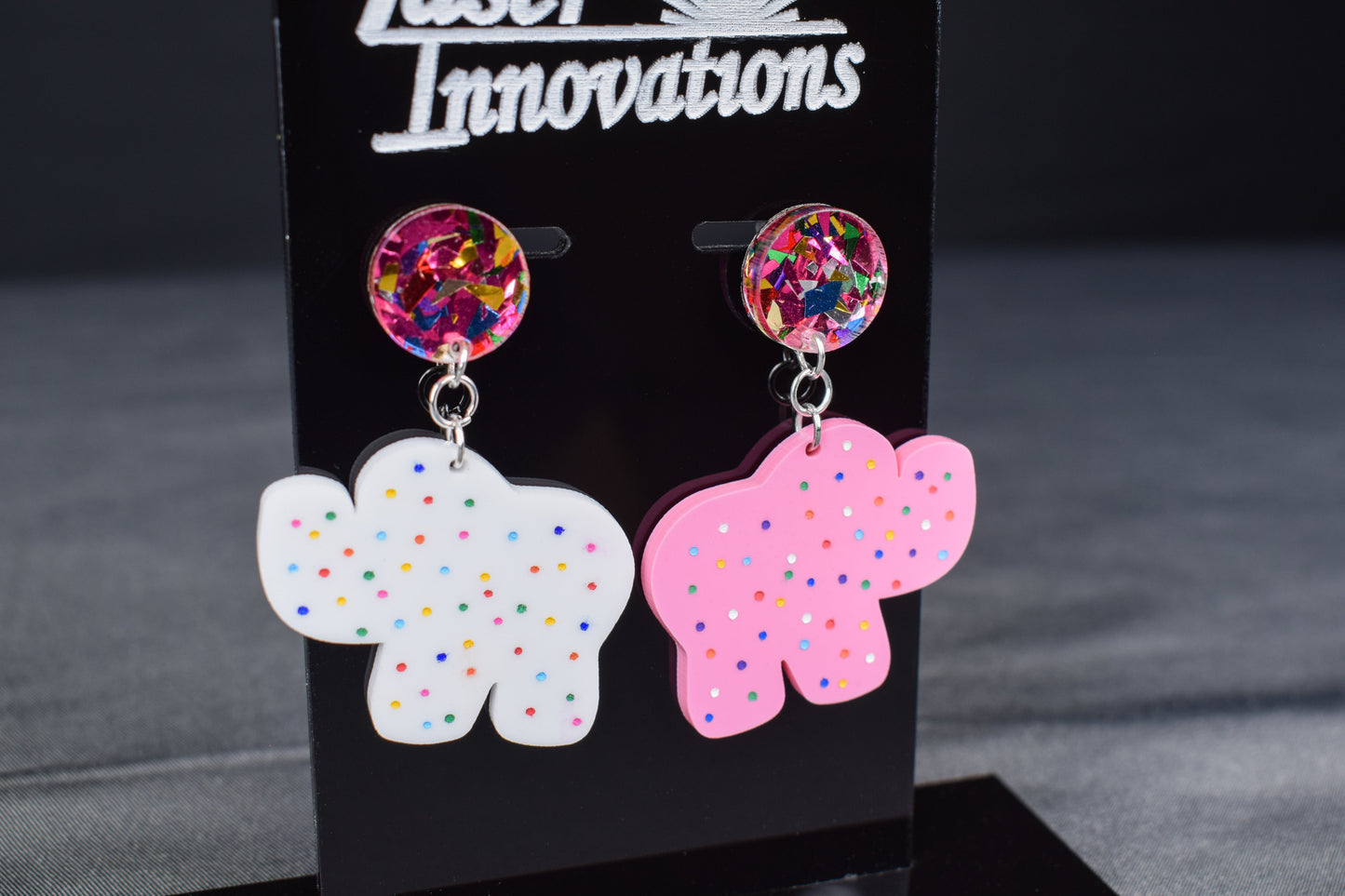 A pair of earrings featuring a white acrylic cookie-shaped elephant and a pink acrylic cookie-shaped camel, with a glittery multicolored acrylic circle attached to each and hand-painted dots