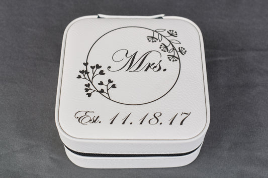 A white synthetic leatherette bridal travel jewelry box with 'Mrs.' and a wedding date laser engraved on the top, laying on a gray surface