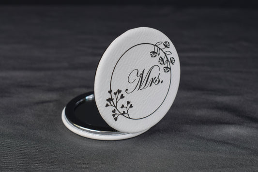 a compact mirror with a white leatherette cover partially opened to display a laser engraving of 'mrs' and a floral design