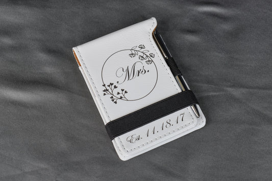A personalized white leatherette bridal vow notebook with custom laser engraving, featuring a floral design and the text 'Mrs' on the cover, resting on a gray surface.