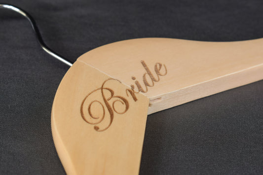 A close up of a wooden hanger with 'bride' laser engraved on it, laying on a dark surface
