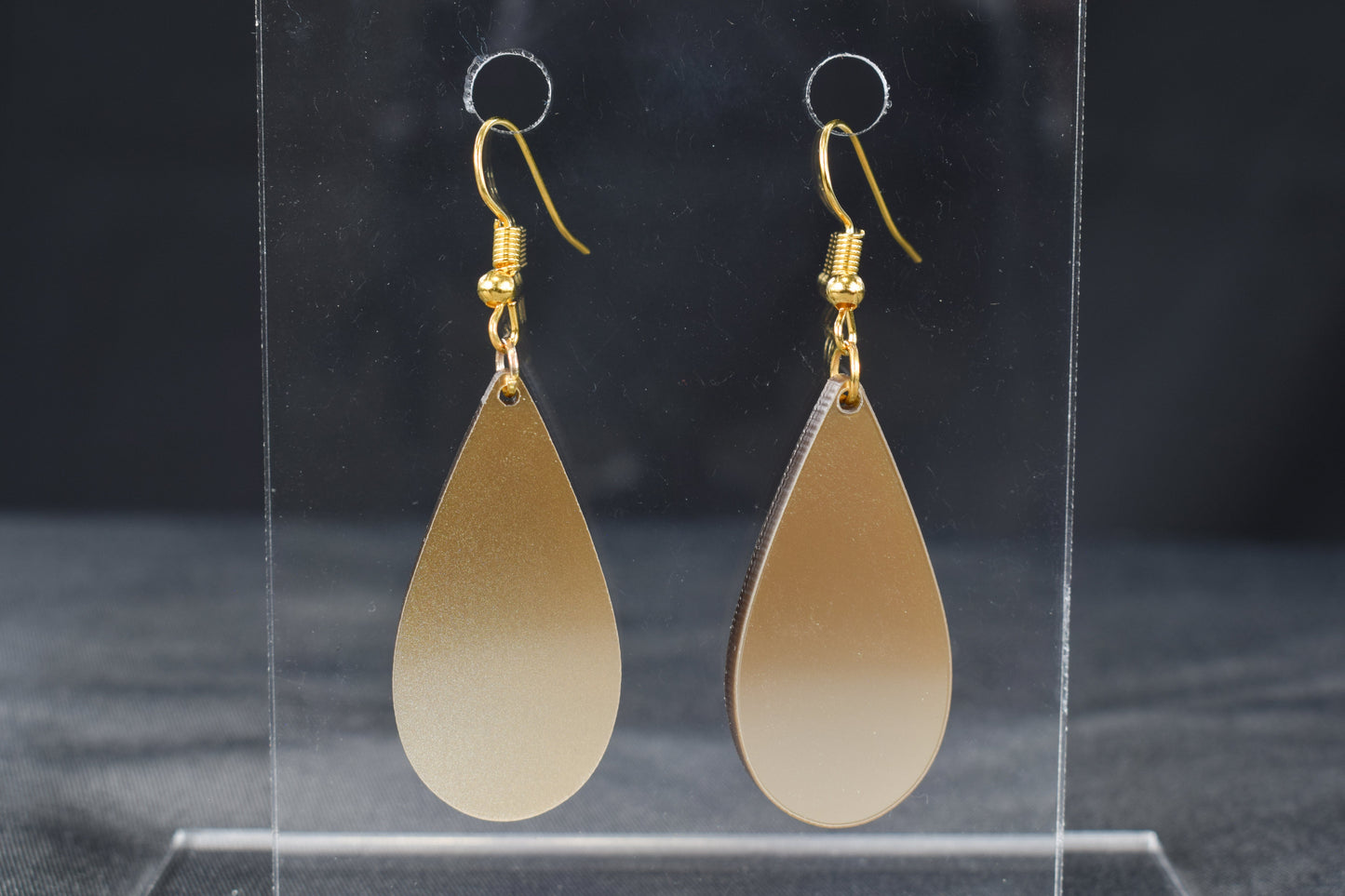A pair of champagne gold metallic acrylic teardrop earrings with gold-plated earring hooks displayed on a clear stand against a black background.