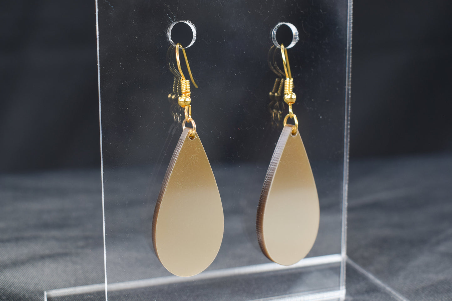 A pair of champagne gold metallic acrylic teardrop earrings with gold-plated earring hooks displayed against a black background.
