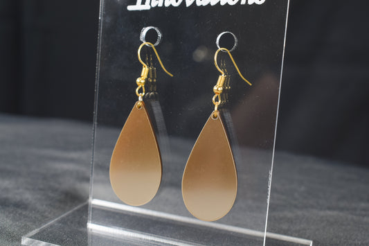 A pair of champagne gold metallic acrylic teardrop earrings with gold-plated earring hooks displayed on a clear stand against a black background.
