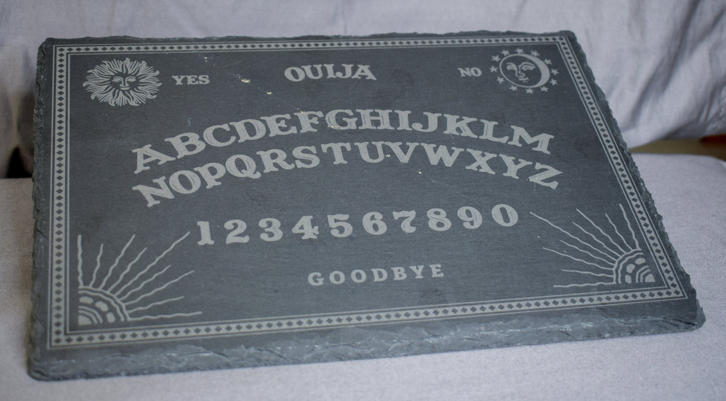 An angled view of a black slate charcuterie board with a Ouija board engraving featuring letters, numbers, and designs, lying on a grey surface.
