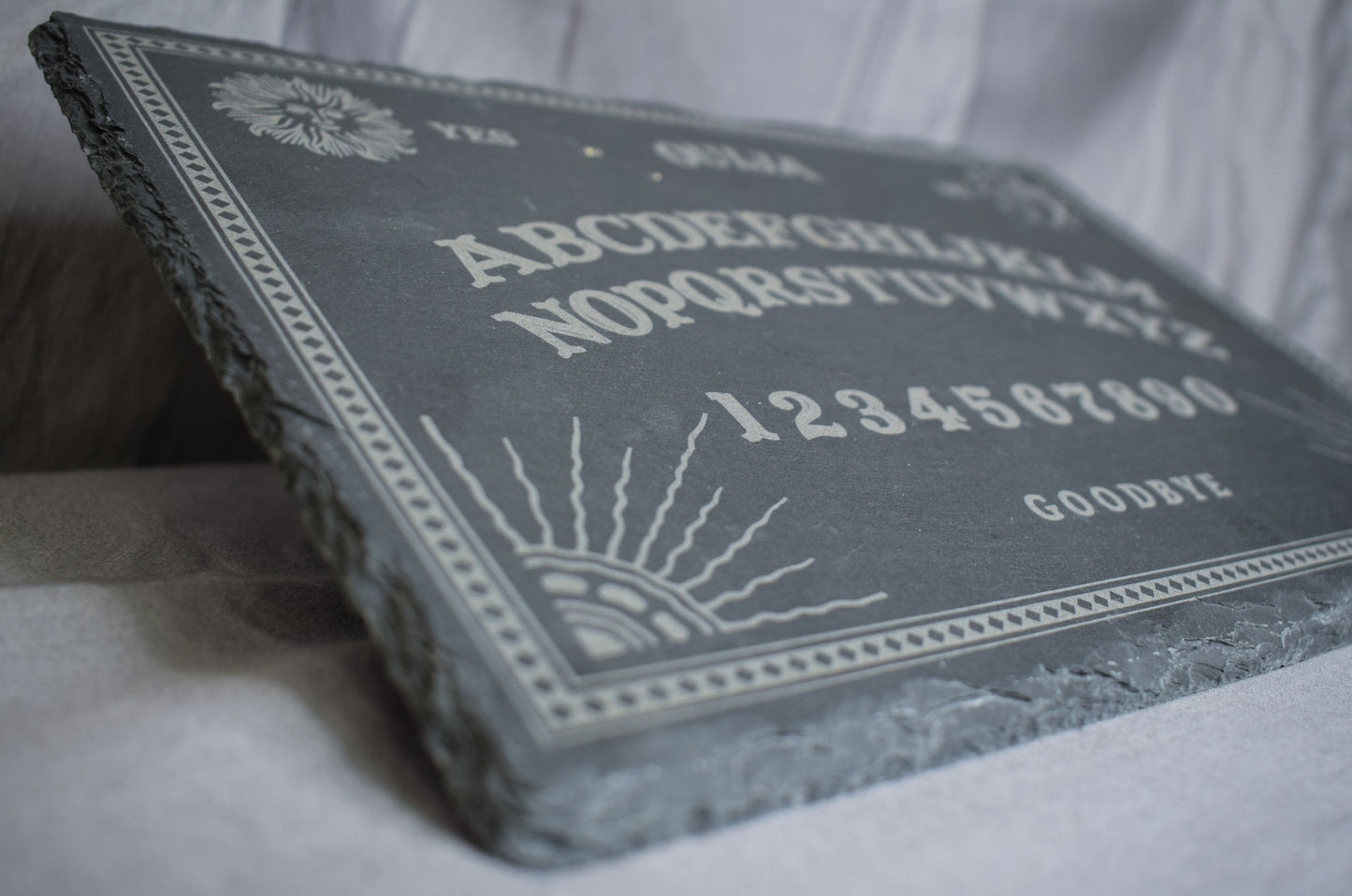 A low angled view of a black slate charcuterie board with a Ouija board engraving featuring letters, numbers, and designs, lying on a grey surface.