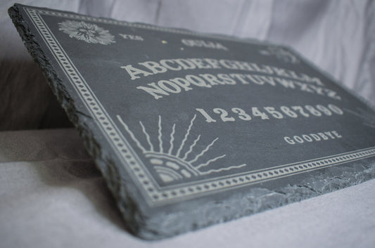A low angled view of a black slate charcuterie board with a Ouija board engraving featuring letters, numbers, and designs, lying on a grey surface.