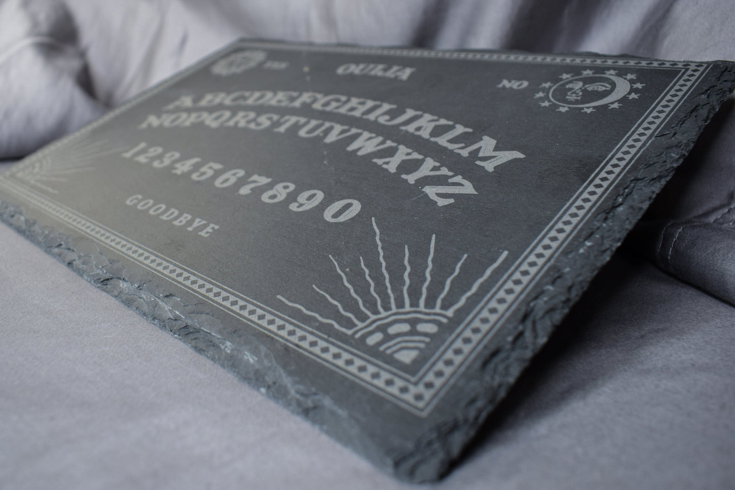 A close up view of a black slate charcuterie board with a Ouija board engraving featuring letters, numbers, and designs, lying on a grey surface.