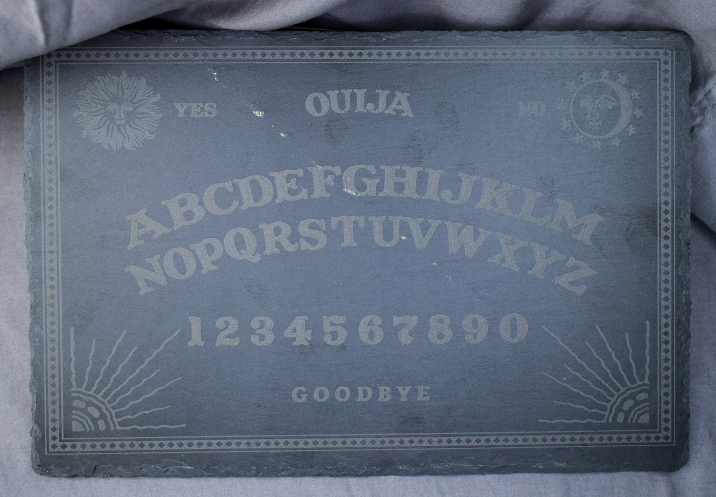 A black slate charcuterie board with a Ouija board engraving featuring letters, numbers, and designs, lying on a grey surface.