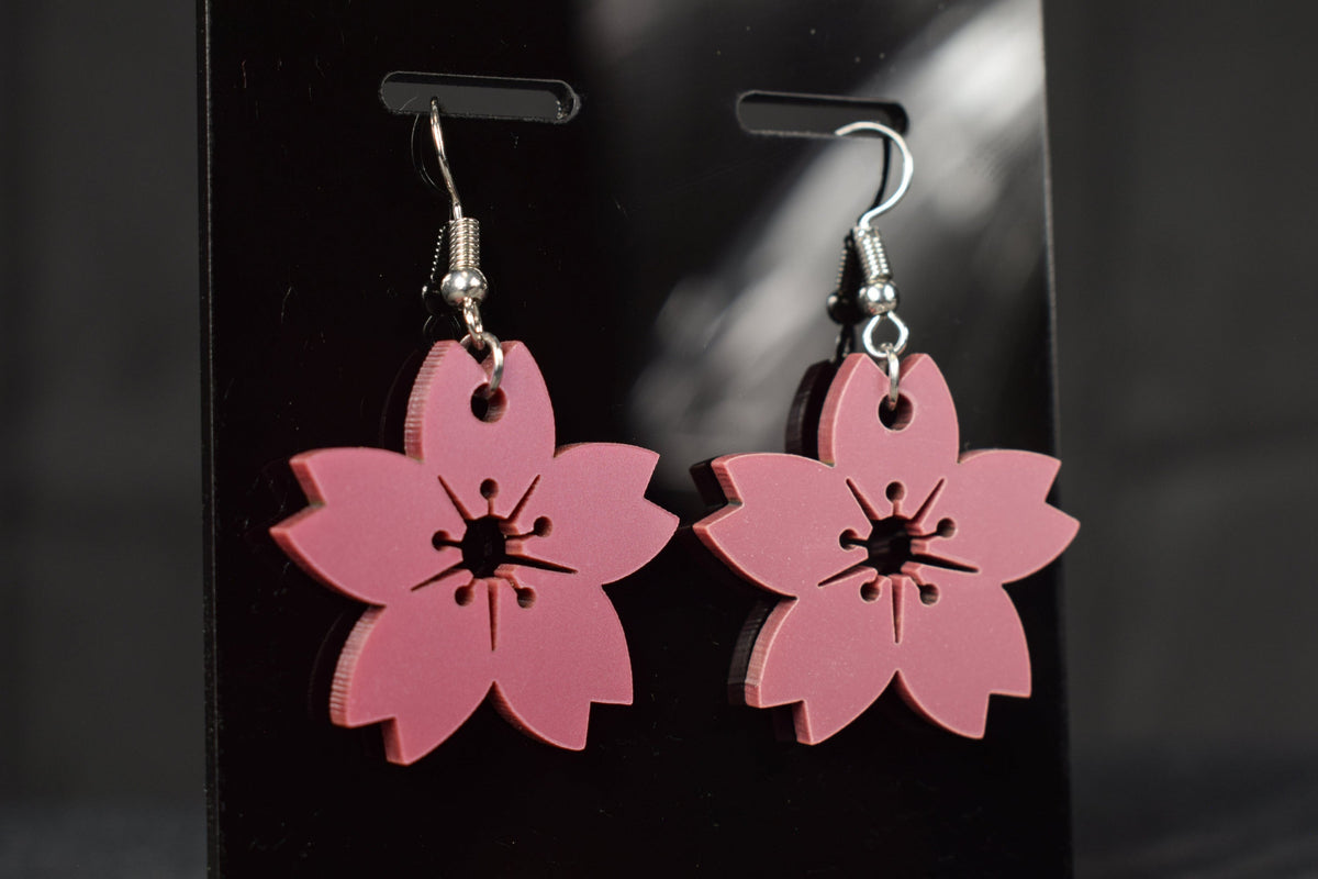 A pair of mauve metallic cherry blossom earrings with silver hooks, displayed on a black stand against a blurred background