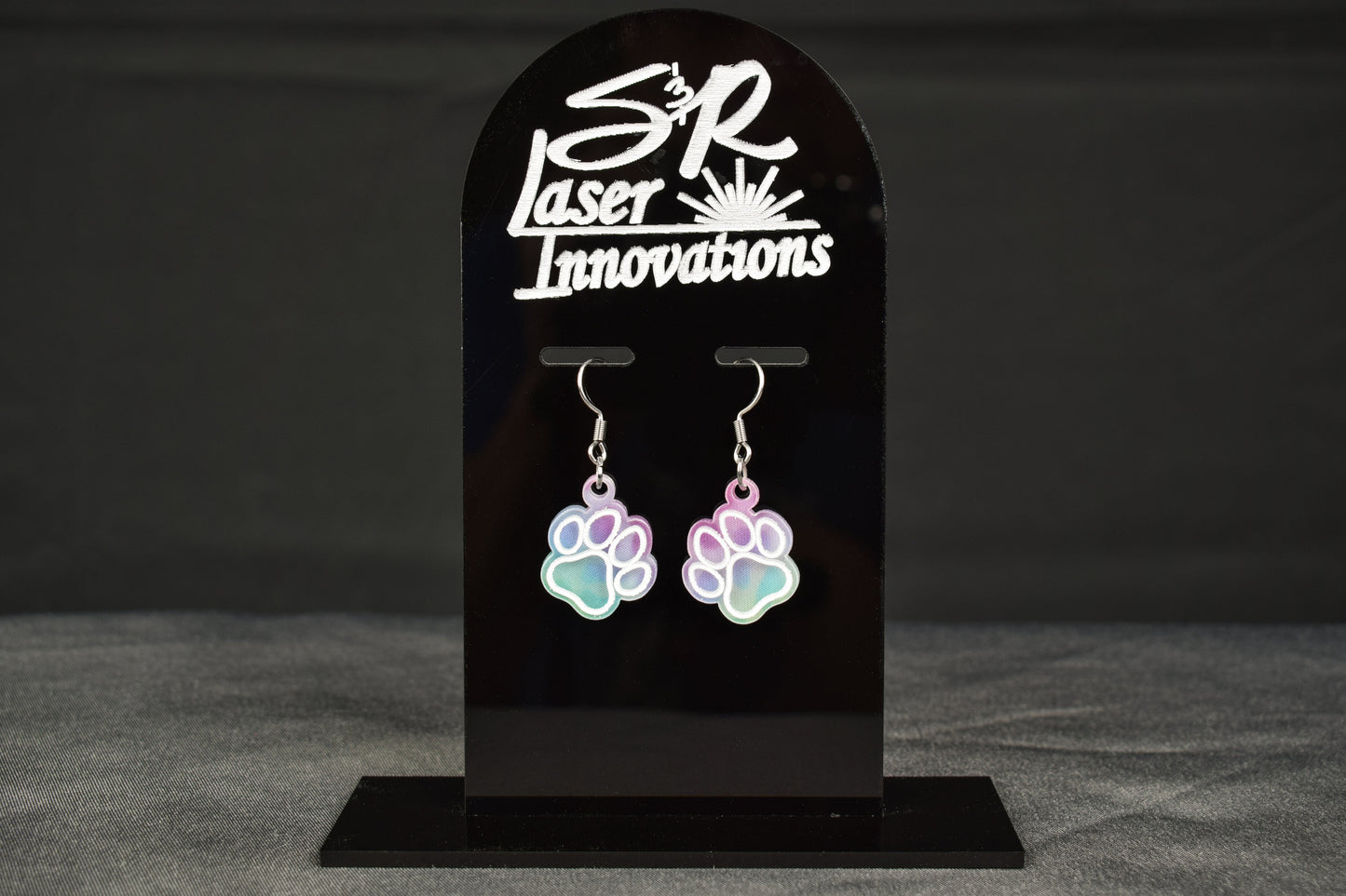 A pair of multicolored earrings with a paw print design and a cool hue, displayed on a stand with 'S&R LASER INNOVATIONS' engraved at the top.