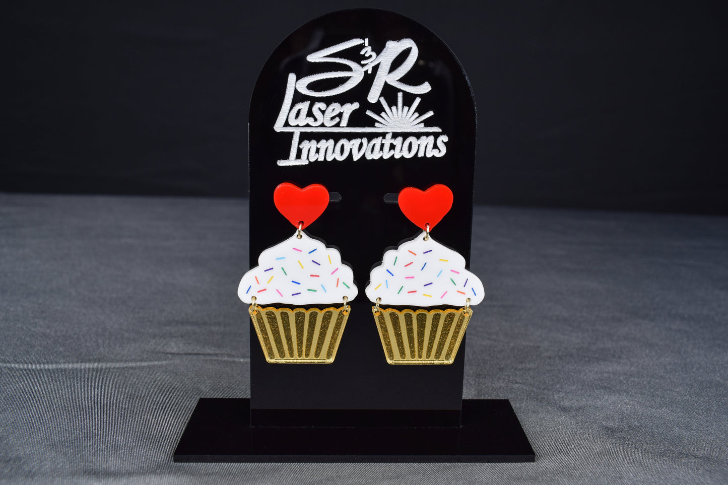 A pair of cupcake-shaped earrings with white acrylic, gold mirror, and red hearts displayed on a black stand with 'SR Laser Innovations' logo.