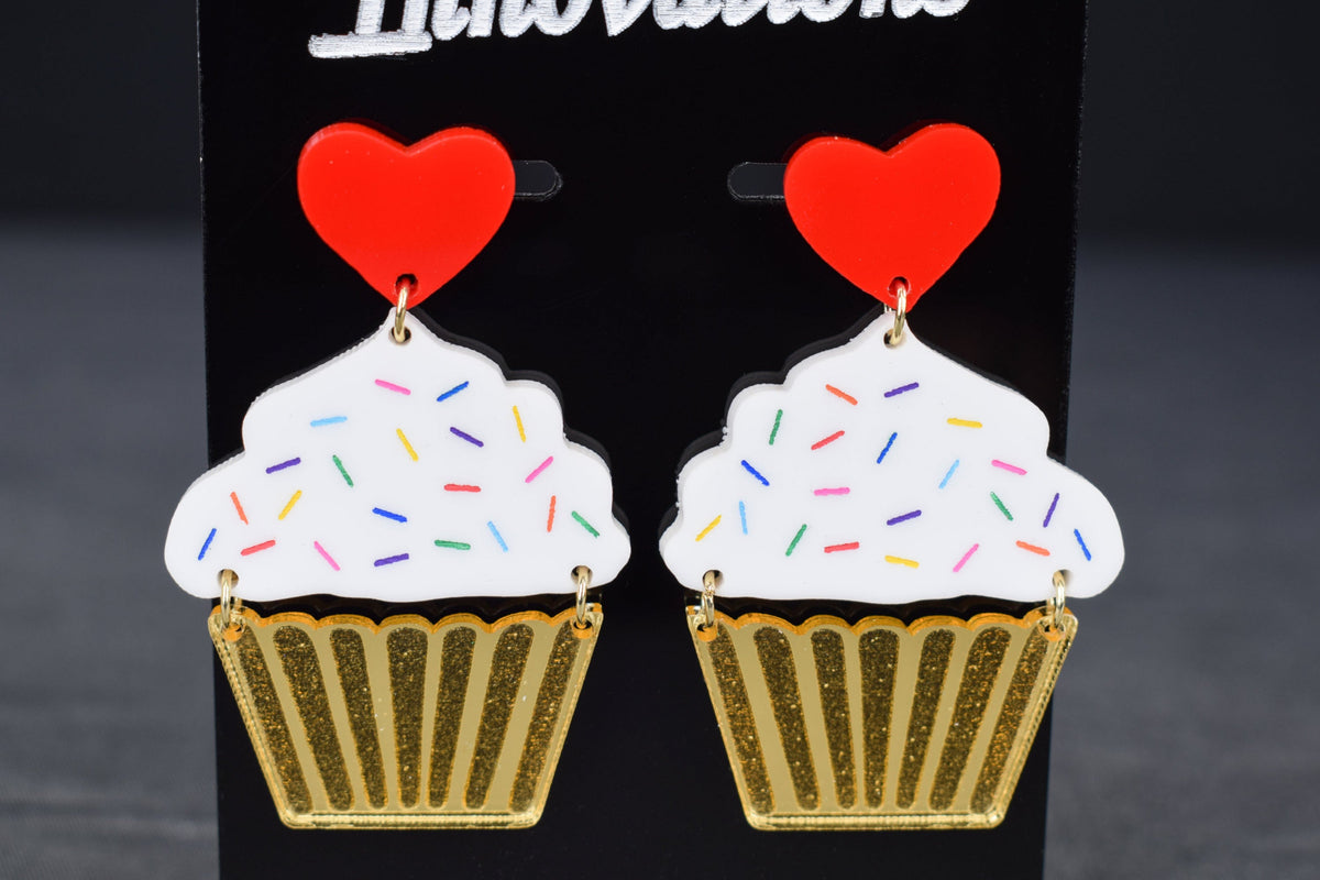 A pair of cupcake-shaped earrings with white acrylic, gold mirror, and red hearts displayed on a black stand