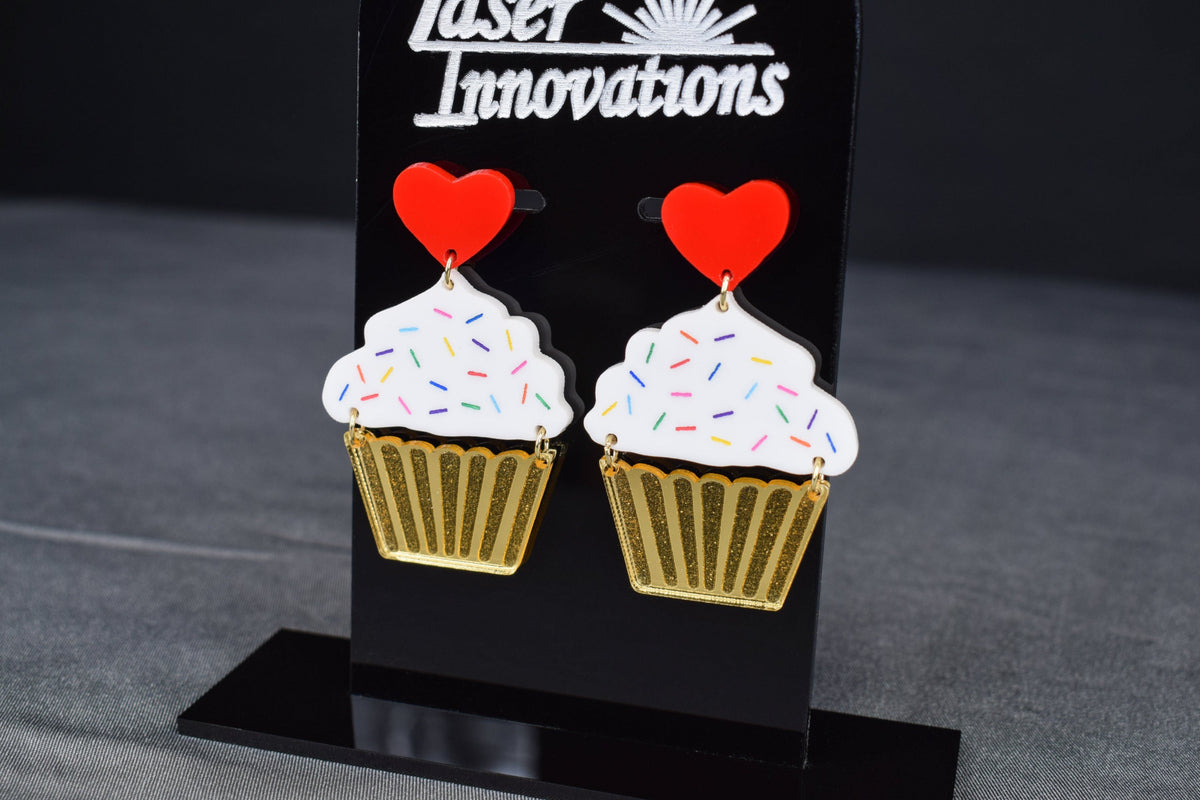 A pair of cupcake-shaped earrings with white acrylic, gold mirror, and red hearts displayed on a black stand with 'SR Laser Innovations' logo.