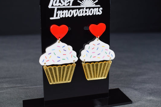 A pair of cupcake-shaped earrings with white acrylic, gold mirror, and red hearts displayed on a black stand with 'SR Laser Innovations' logo.