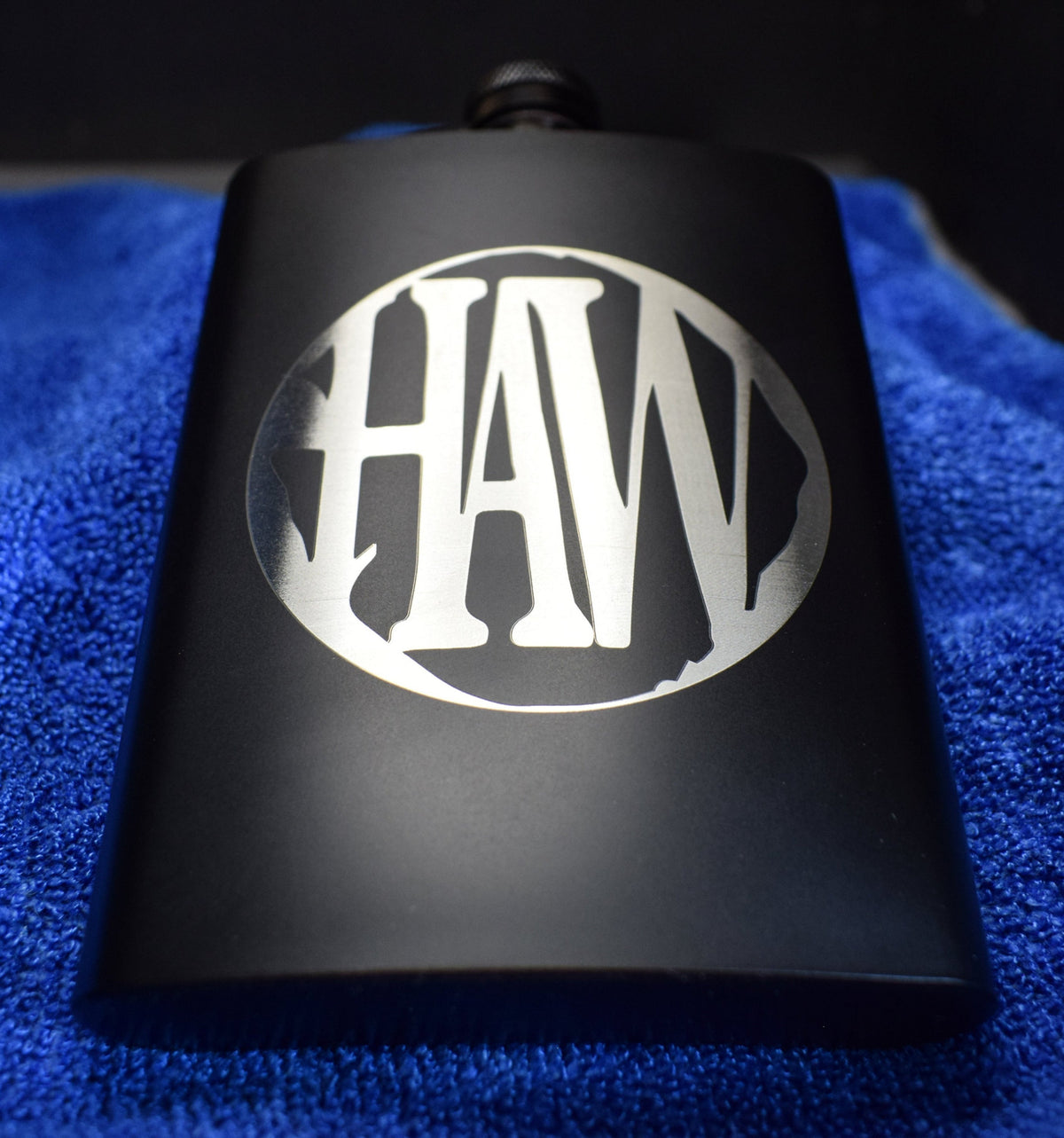 A black metal flask with a custom engraving of the logo 'HAW' on a blue surface.
