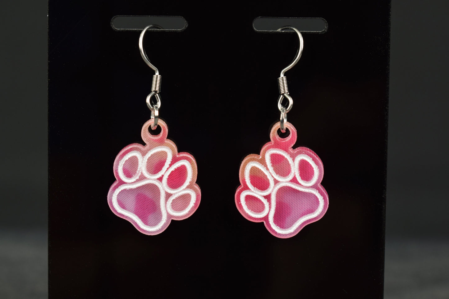 A close up of colorful acrylic paw print earrings with a bright hue, displayed on a black stand
