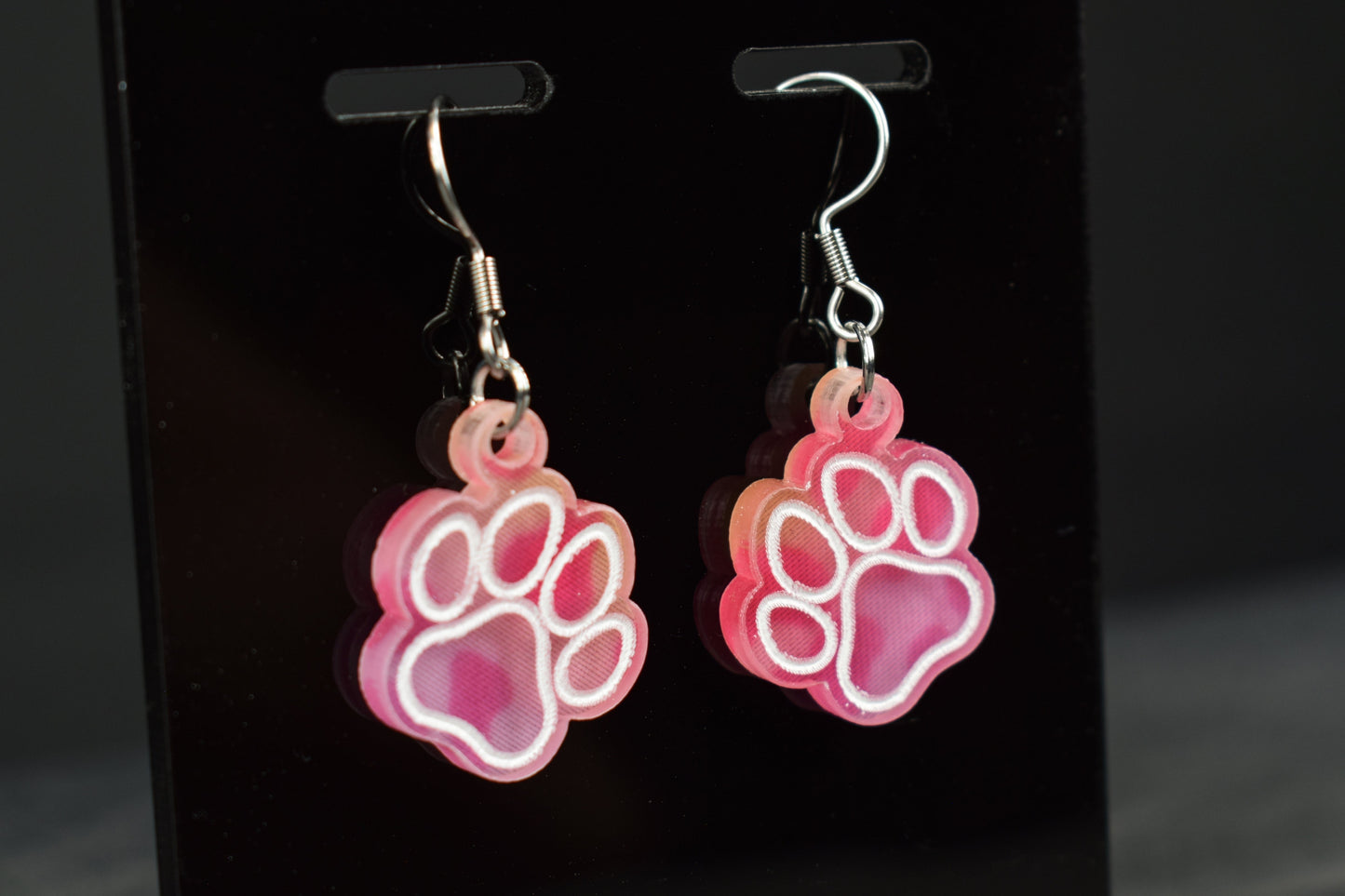 A close up of colorful acrylic paw print earrings with a bright hue, displayed on a black stand