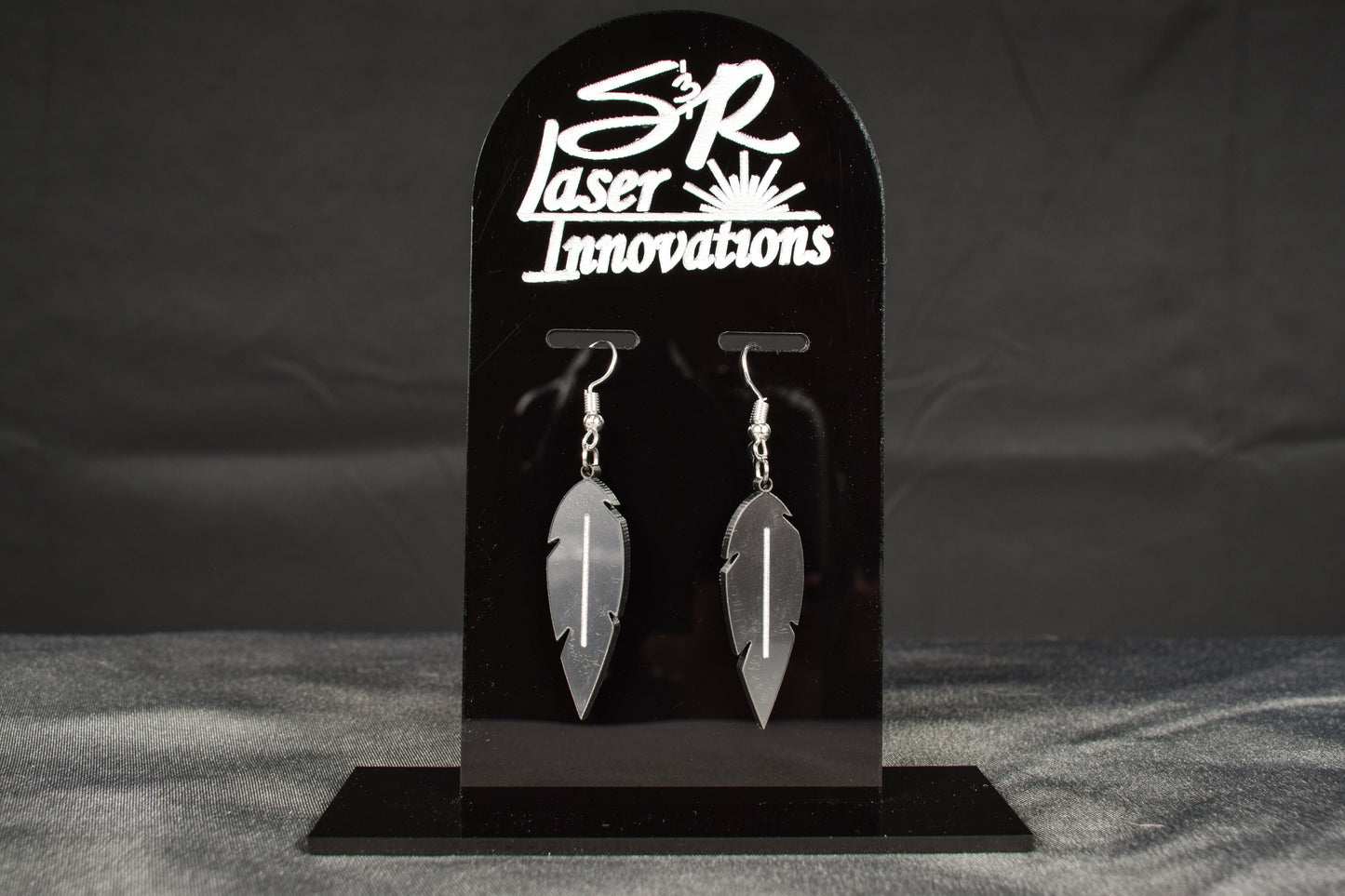 A pair of charcoal metallic acrylic feather earrings displayed on a black stand with 'SR Laser Innovations' branding above.
