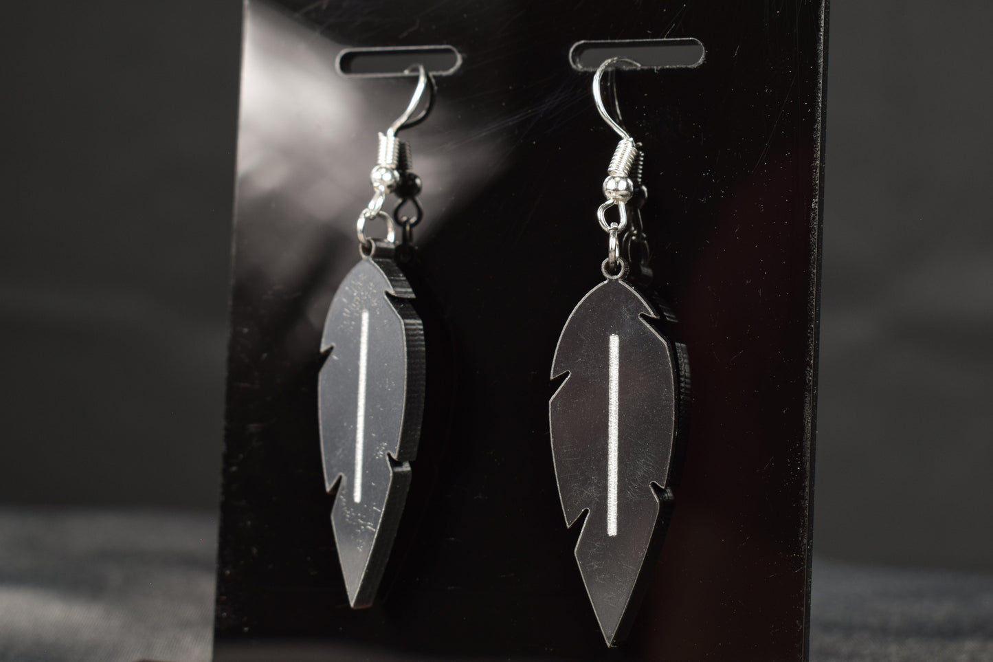 A close up view of a pair of charcoal metallic acrylic feather earrings displayed on a black stand, showcasing the silver hooks and white stripe in the middle of the earrings