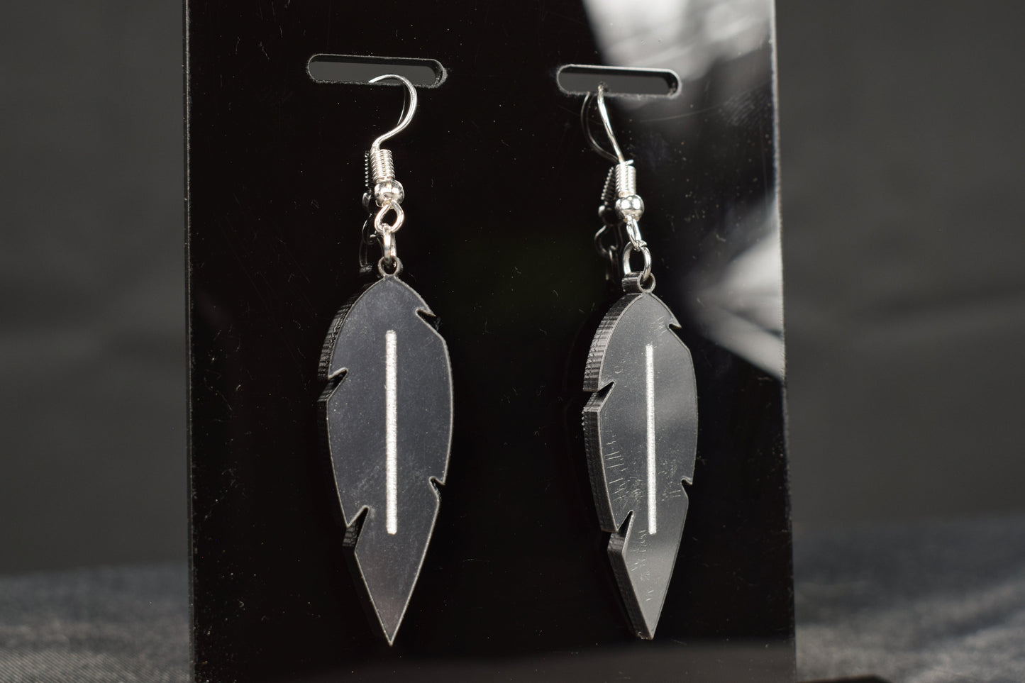 A close up view of a pair of charcoal metallic acrylic feather earrings displayed on a black stand, showcasing the silver hooks and white accent stripe