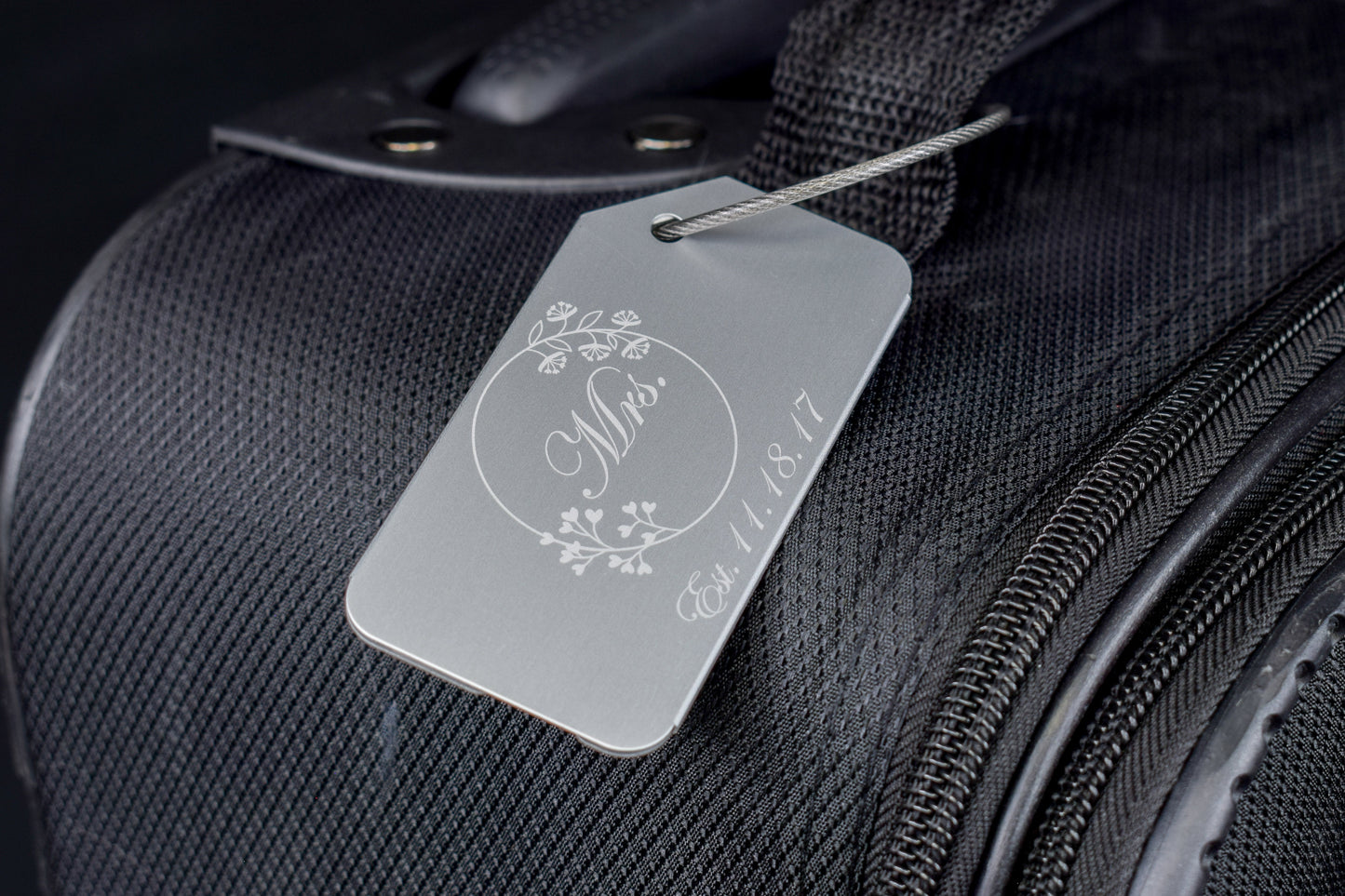 a silver aluminum luggage tag with a white laser engraving of 'Mrs' and a floral design, attached to black luggage