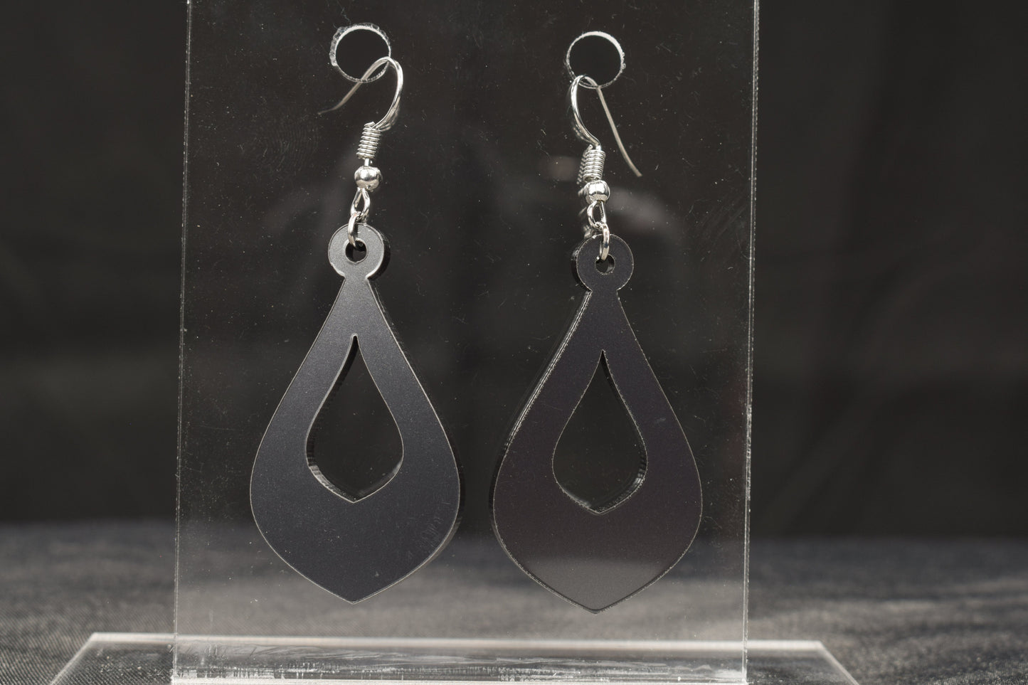 a pair of teardrop shaped hoop style earrings in charcoal metallic acrylic displayed on a clear stand, featuring stainless steel hooks
