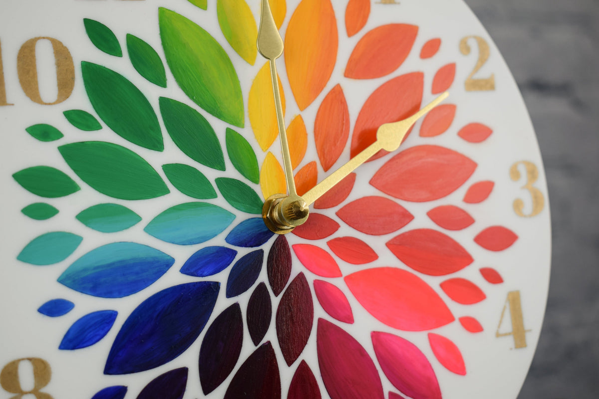 A close up view of a hand painted rainbow clock, showcasing the flower petals and gold hands, set against a blurred background