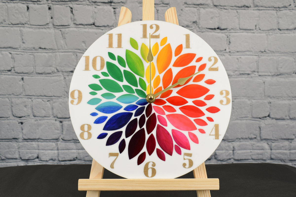 A round, hand-painted acrylic clock with a rainbow flower design, displayed on a wooden easel, set against a brick wall background.