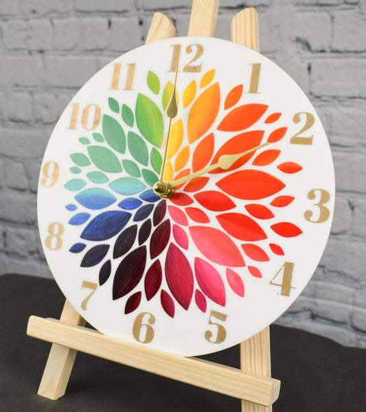 A hand painted clock with rainbow colored flower petals and gold hour numbers, displayed on an easel against a white brick background