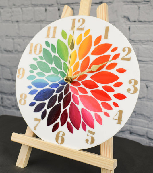 A hand painted clock with rainbow colored flower petals and gold hour numbers, displayed on an easel against a white brick background