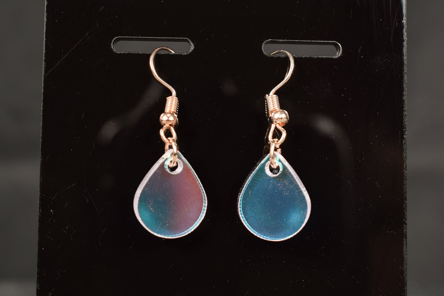 A close up view of a pair of iridescent rainbow teardrop earrings displayed on a black stand