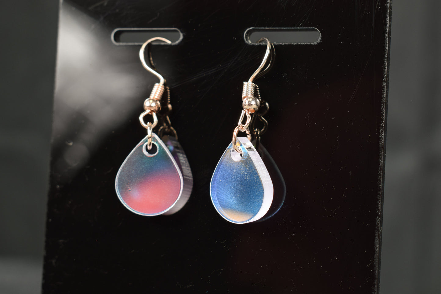 A close up view of a pair of iridescent rainbow teardrop earrings displayed on a black stand