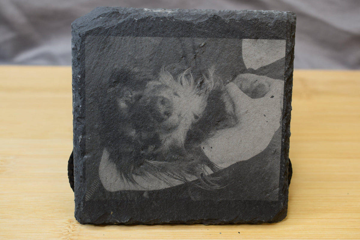 A black slate square coaster with an image of a dog laser engraved on the surface, displayed on a wooden surface