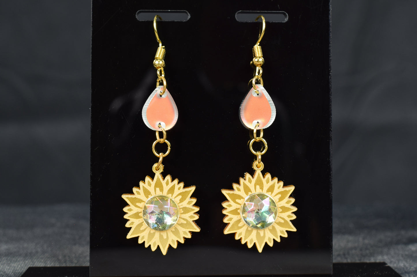 A pair of gold metallic and iridescent acrylic sunflower drop earrings displayed on a black stand