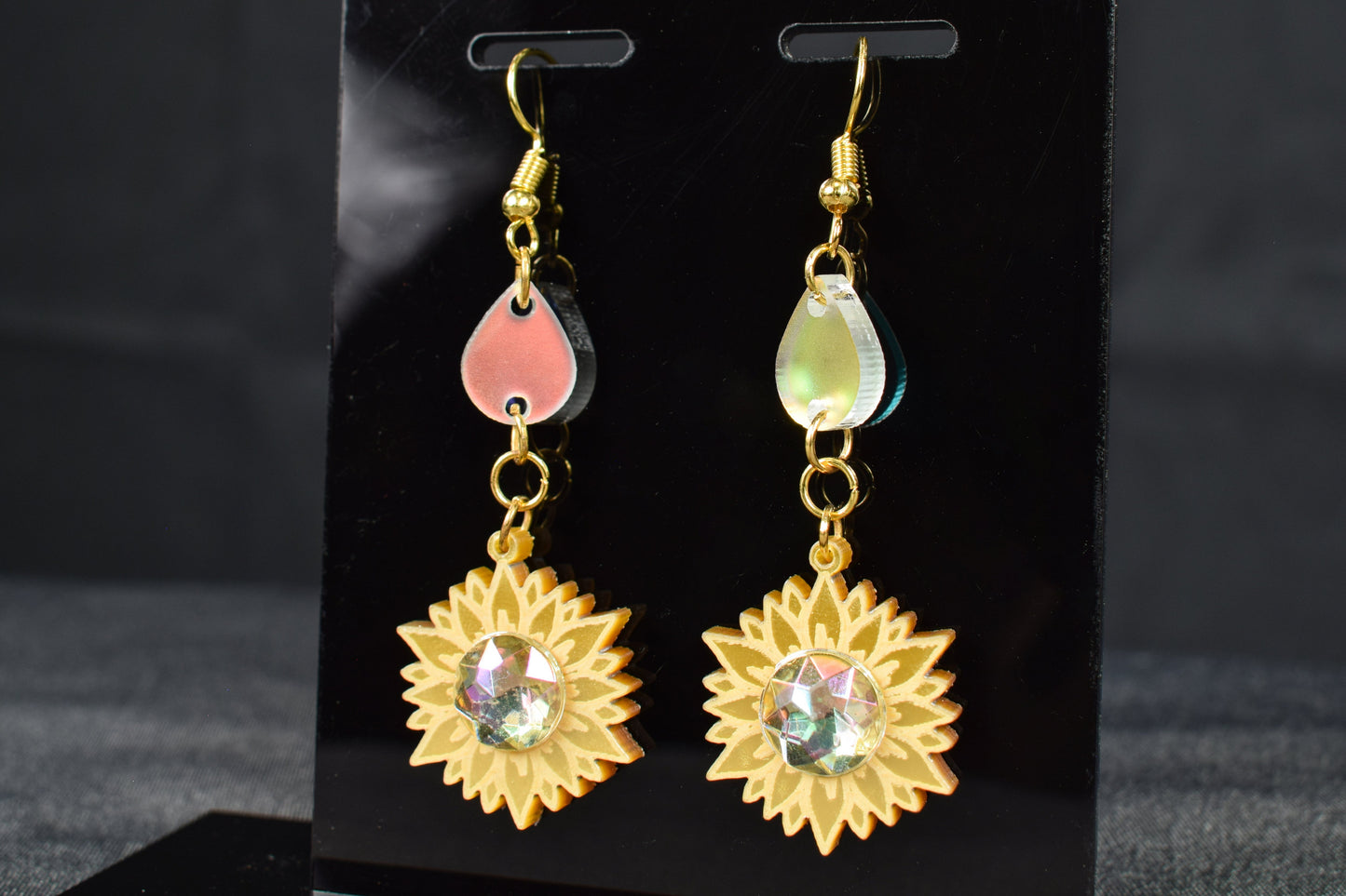 A pair of gold metallic and iridescent acrylic sunflower drop earrings displayed on a black stand