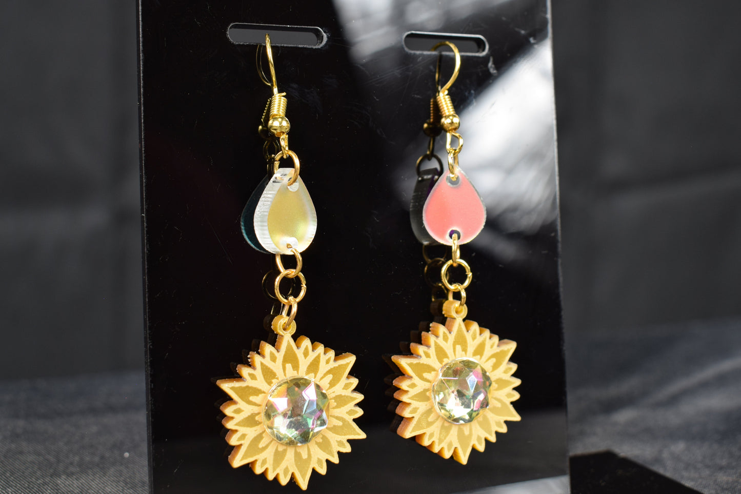 A pair of gold metallic and iridescent acrylic sunflower drop earrings displayed on a black stand