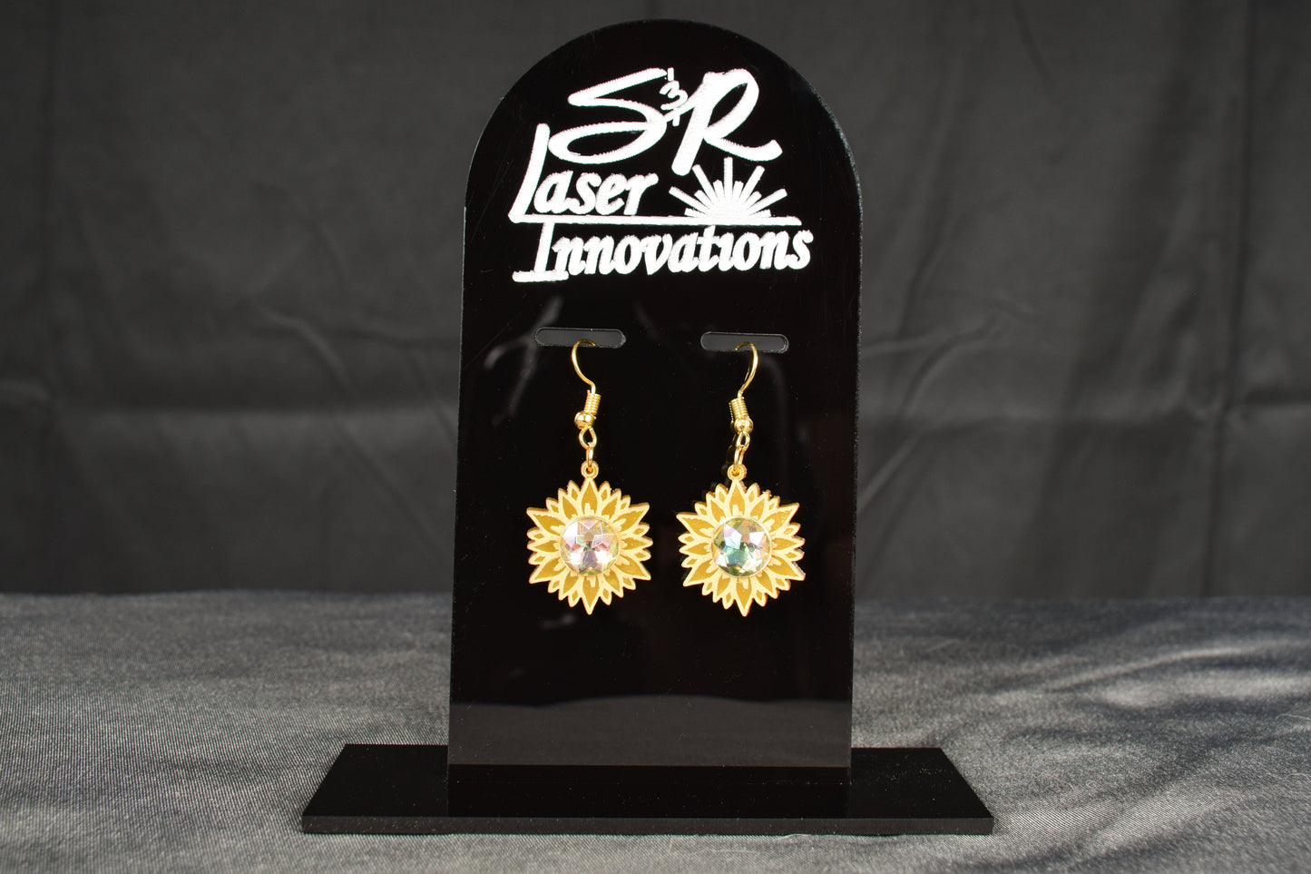 A pair of gold metallic acrylic earrings designed to resemble sunflowers with iridescent rhinestone centers, displayed on a black stand with 'SR Laser Innovations' branding.