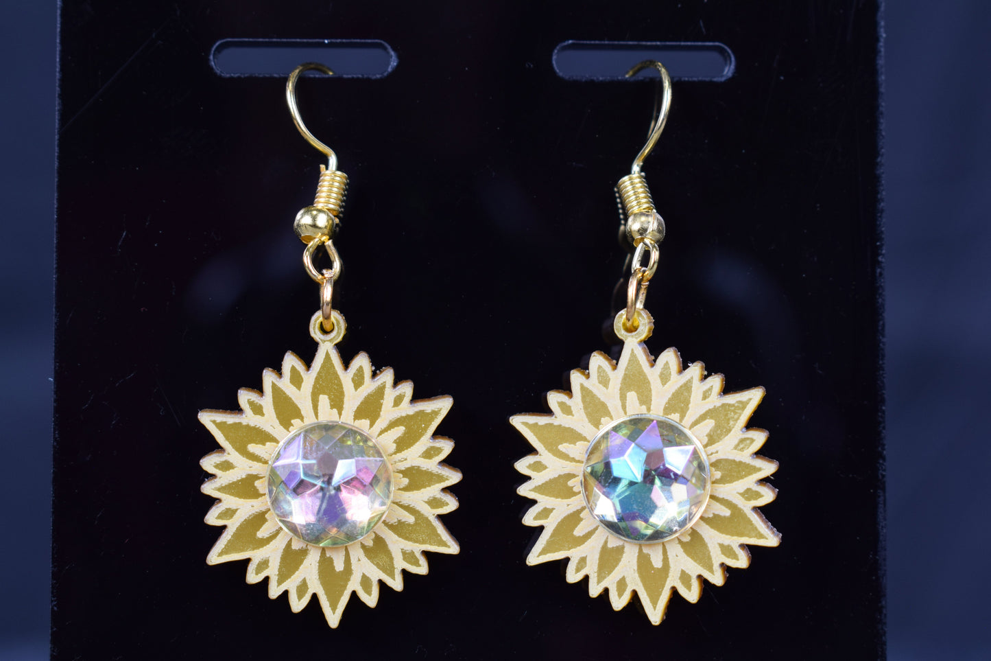 A close up of a pair of gold metallic acrylic earrings designed to resemble sunflowers with iridescent rhinestone centers, displayed on a black stand