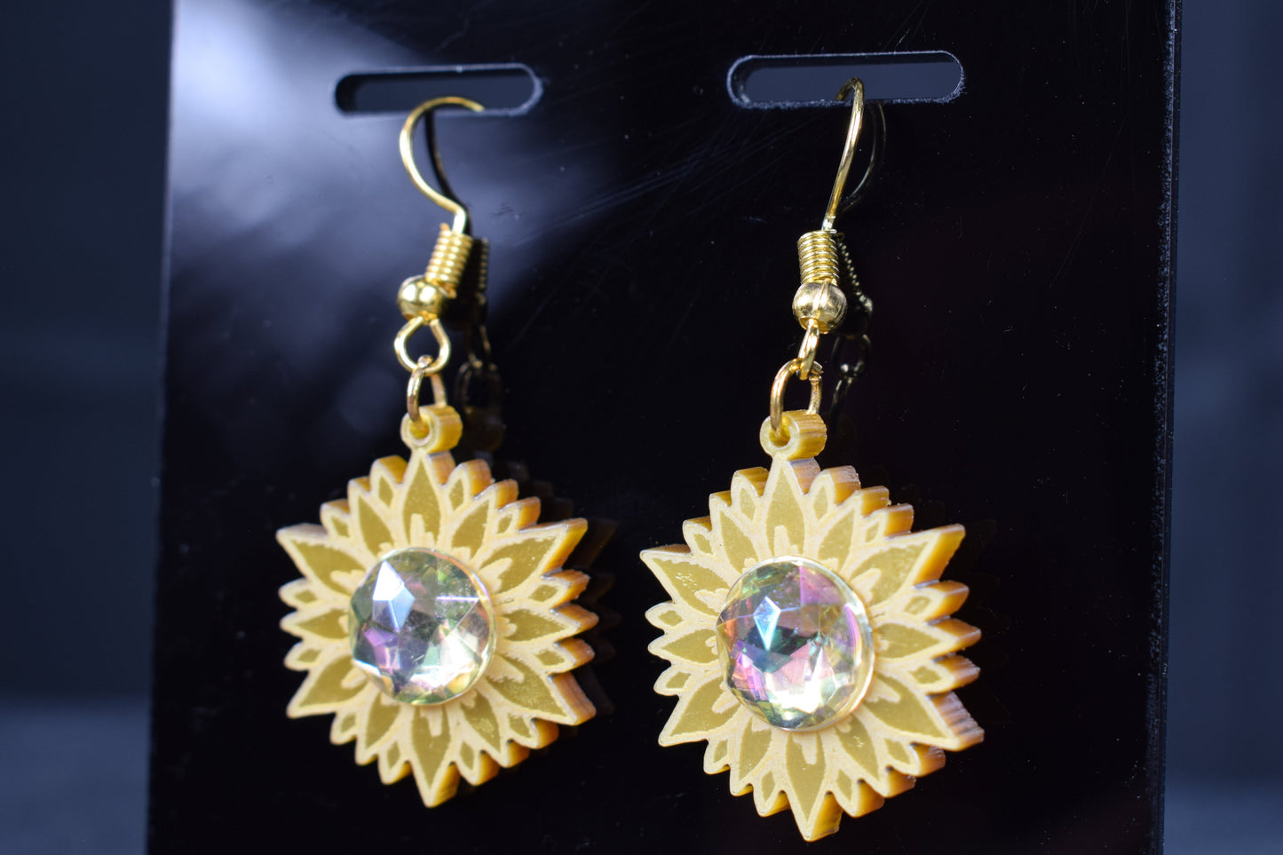 A close up of a pair of gold metallic acrylic earrings designed to resemble sunflowers with iridescent rhinestone centers, displayed on a black stand with 'SR Laser Innovations' branding.