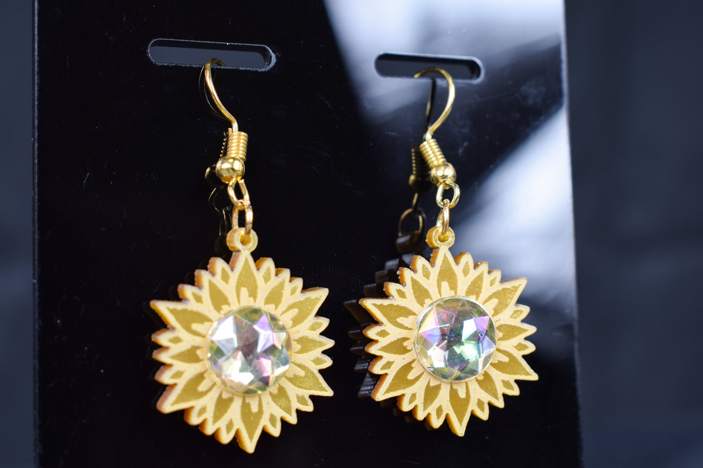 A close up of a pair of gold metallic acrylic earrings designed to resemble sunflowers with iridescent rhinestone centers, displayed on a black stand