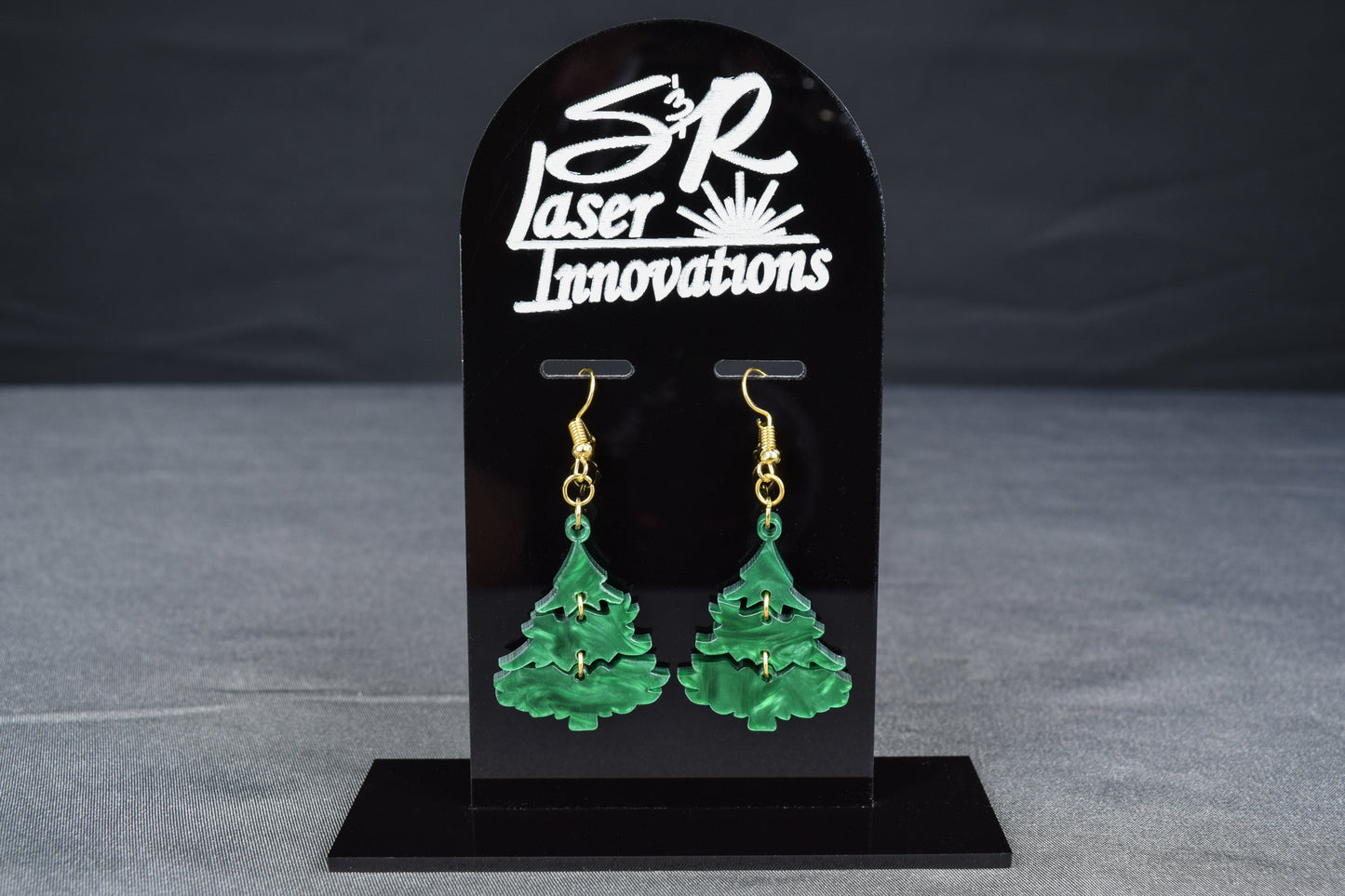 A pair of three-tiered Christmas tree earrings made of dark green swirl acrylic, displayed on a black stand with 'SR Laser Innovations' branding.