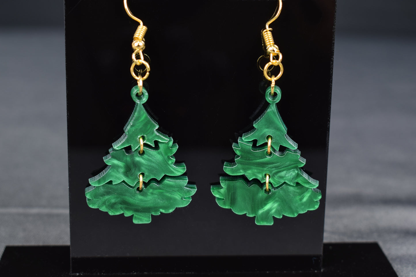 A pair of three-tiered Christmas tree earrings made of dark green swirl acrylic, displayed on a black stand with 'SR Laser Innovations' branding.