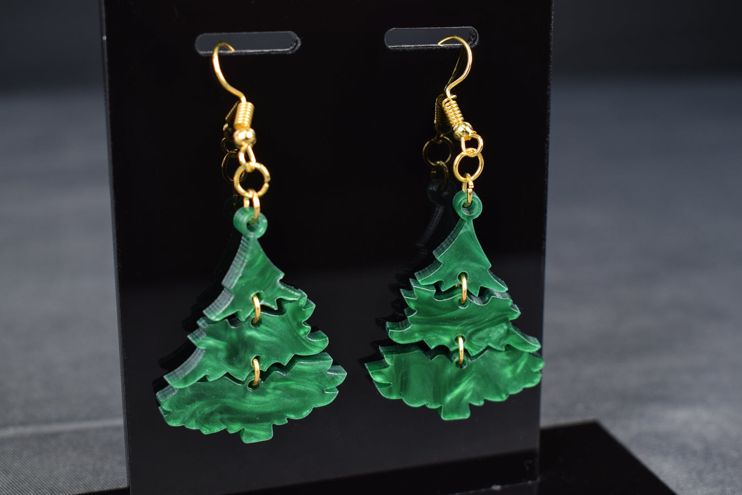 A pair of three-tiered Christmas tree earrings made of dark green swirl acrylic, displayed on a black stand with 'SR Laser Innovations' branding.