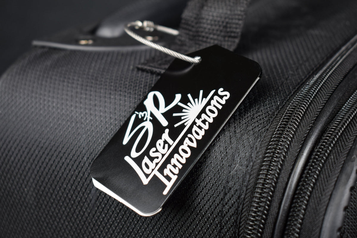 A black aluminum luggage tag with custom logo engraving, attached to a piece of luggage.