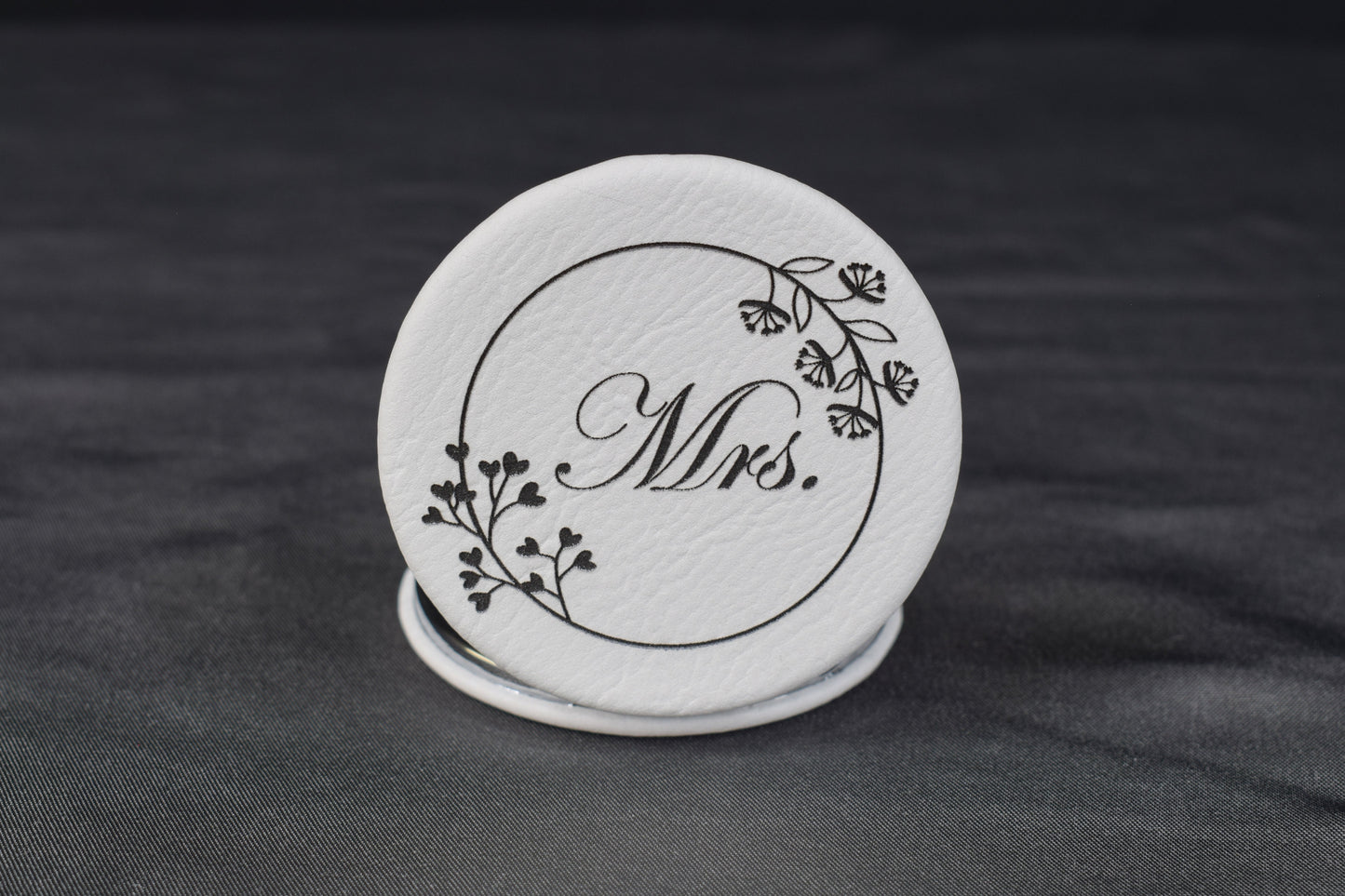 A round compact mirror with a white synthetic leatherette cover, featuring a laser-engraved design with the text 'Mrs.' and floral elements.
