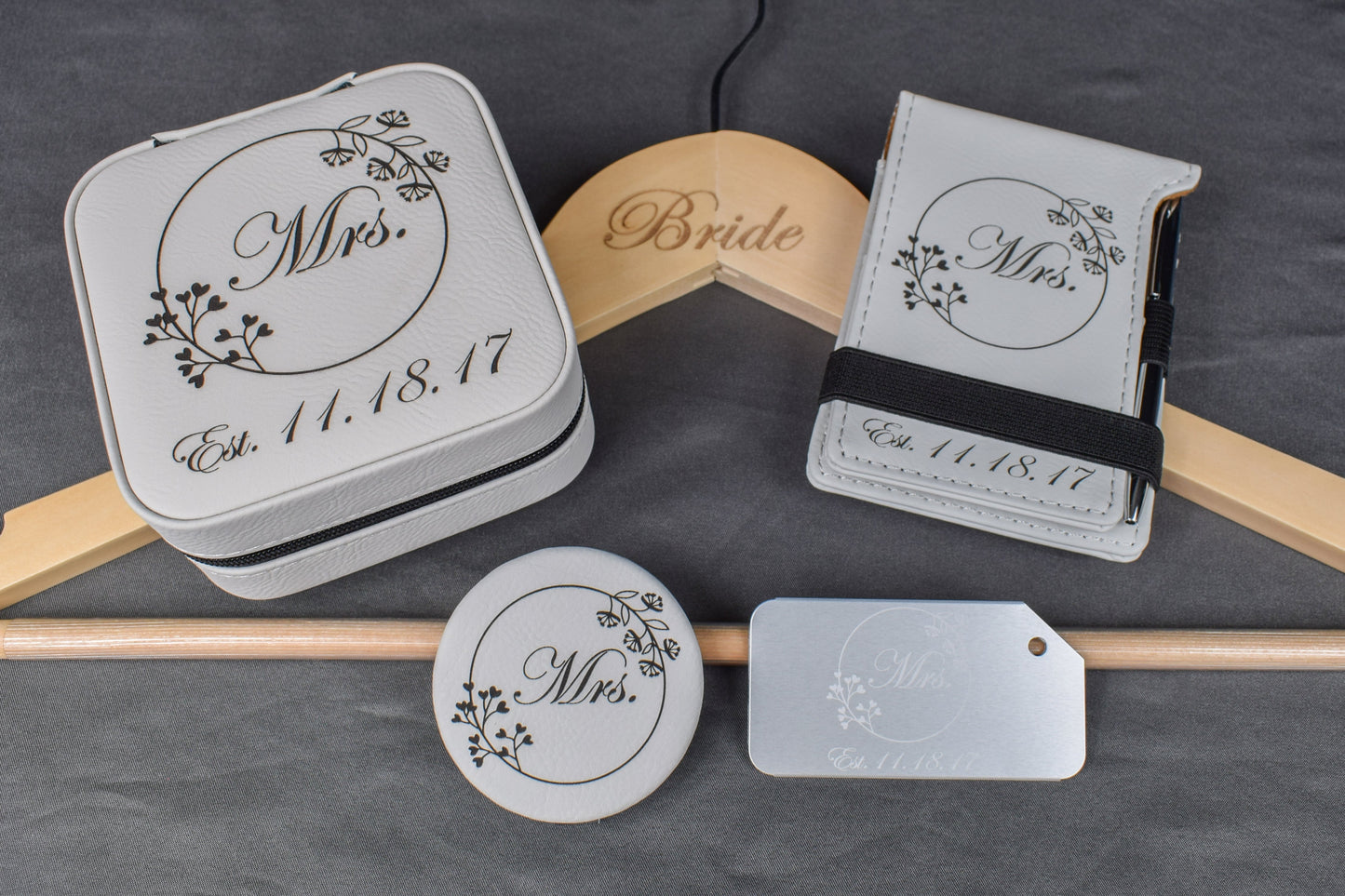 A set of personalized bridal gift items including a custom aluminum luggage tag, a white compact mirror, a white vow mini notebook, a custom engraved bridal hanger, and a custom laser engraved travel jewelry box, all with elegant designs and the wedding date.