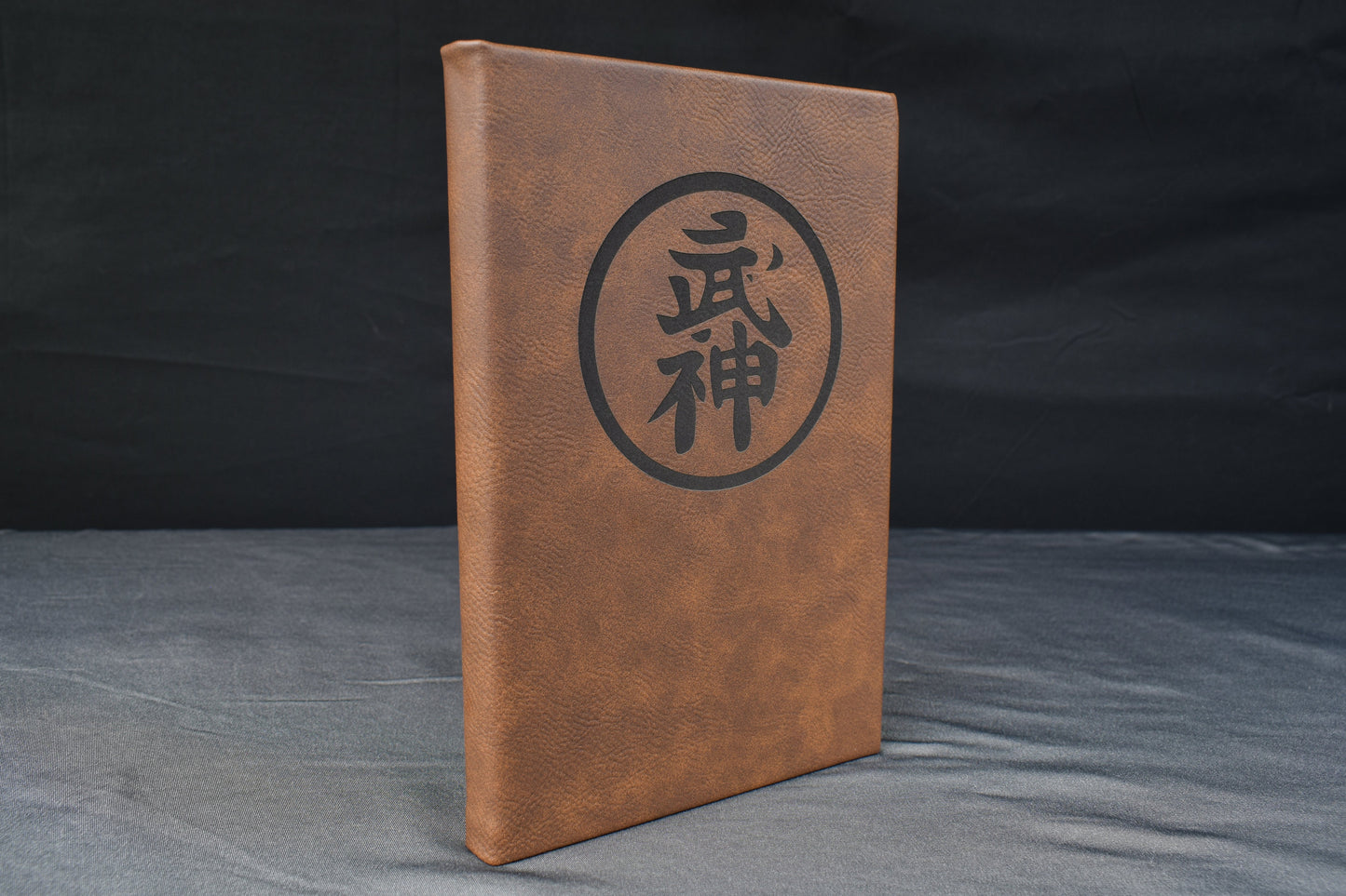 a brown leatherette journal with a prominent symbol laser engraved on the cover, against a black background