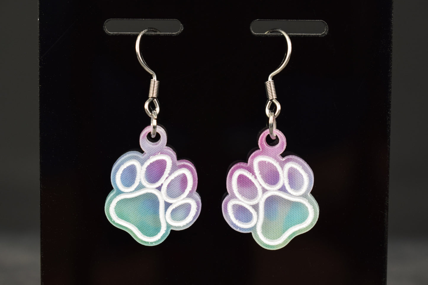 A close up of a pair of multicolored earrings with a paw print design and a cool hue, displayed on a black stand